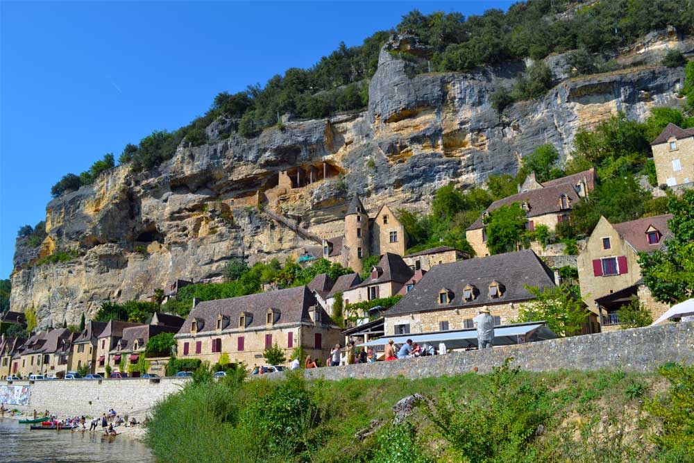 Fransen houden van labels en van afkortingen en met PBVF weten ze beide aardig te combineren. PBVF staat voor Plus Beaux Villages de France, de Mooiste Dorpen van Frankrijk. Nu telt Frankrijk natuurlijk ontzettend veel gemeenten. 35.416 om precies te zijn. Ze zijn vaak niet groot en tellen gemiddeld slechts weinig inwoners. Veel van die dorpjes zijn best mooi, maar 164 ervan zijn bestempeld als wondermooi en kregen het label. En niet minder dan 13 daarvan staan op het programma van onze gasten. Dat wordt keuzes maken! Welke dorpen kunnen PBVF worden? Om Plus Beaux Village de France te worden dien je als dorp een aanvraag in. Let wel, de criteria zijn bijzonder streng: slechts 20% van de aanvragers ontvangt na doorgedreven evaluatie het begeerde label. Er zijn dan ook niet minder dan 30 criteria. Om te weten of het überhaupt de moeite loont om een aanvraag in te dienen kijk je best zorgvuldig naar de eerste drie criteria: je dorp telt niet meer dan 2000 inwoners, je botst er op ten minste twee beschermde sites, zoals een historisch monument of een opmerkelijke erfgoedlocatie én de aanvraag wordt gedragen door de gemeenteraad. Check? Ga dan door naar de volgende ronde. Welke selectiecriteria hanteert de commissie van de PBVF? Vooreerst wordt er gekeken naar de beschermde sites en monumenten in de dorpskern. Dat moeten er dus minstens twee zijn, maar meer mag altijd. Dan worden de architectonische kwaliteiten van het dorp als geheel onder de loep genomen. Harmonie en homogeniteit, daar draait het om. Passen de gebouwde volumes mooi bij elkaar? Is er uniformiteit in de dakbedekkingsmaterialen, de kleuren van de daken, de gevelmaterialen en de gevelkleuren? Mooi. Dan kijken we verder naar de stedenbouwkundige kwaliteiten. Hoe omvangrijk dan wel compact is het bebouwde weefsel? Is ook de omgeving en de toegang tot het dorp de moeite waard? En hoe zit het met de dimensie van het wegennet? Tot slot gaat de commissie na of je als dorp ook actief de nodige inspanningen levert om het geheel nog bijkomend te verfraaien. Wat is het gevoerde beleid aangaande renovatie van gebouwen, reclamepanelen, inrichting van de openbare ruimte, inbreng en beheer van groenelementen? Hoe organiseer je het parkeren in en om het dorp, wat met elektriciteits- en telefoondraden, hoe esthetisch verantwoord is de openbare verlichting?... Verwacht je dus maar aan een lijvig dossier. Liggen er Plus Beaux Villages in de omgeving van Le Manoir? Jazeker, dertien dus… al dient het gezegd dat we, zoals we het hier op het Franse platteland gewoon zijn, ‘omgeving’ best wel ruim interpreteren. Zo ligt ‘onze’ hoofdstad Cahors, de hoofdstad van het departement Lot, op zo’n 70 km. Dat is nu niet meer meteen een dingetje - onze administratie staat tegenwoordig helemaal op punt - maar de eerste jaren zijn we geregeld heen en weer gereden met formulier x en aanvraagblad y (en of we bewijsstuk z misschien ook nog even kunnen binnenbrengen?). Ook voor andere dan onze dagelijkse boodschappen rijden we al snel de ruim 30 km tot Brive-la-Gaillarde. Een concertje in Gramat? 35 km. Een lezing in Milandes? 41 km. Mét bochten. Maar stuk voor stuk schitterende wegen door een wondermooi landschap. We blijven van elke tocht genieten en onze gasten hebben we ook nog niet horen klagen. Daar gaan we dus, richting PBVF… Een overzicht van wat de commissie ons te vertellen heeft, in alfabetische volgorde: Autoire, in het departement Lot, op 44 km van Le Manoir in Souillac Autoire ligt in de holte van een keteldal: een half cirkelvormige vallei die ooit door een gletsjer zou zijn gevormd. Het keteldal wordt omgeven door de ‘causse’ - het kalkplateau - tussen Figeac en Gramat. Hier veel landhuizen en herenhuizen, met consequent bruine dakpannen en een veelheid aan karakteristieke vierkante duiventillen. Een wandeling richting de wand van het keteldal is een must: tijdens nattere perioden klatert er met veel geweld een waterval naar beneden. Verder wandelen tot in Loubresac (zie verder) kan ook. Beynac-et-Cazenac, in het departement Dordogne, op 35 km van Le Manoir in Souillac Beynac-et-Cazenac bevindt zich op 10 km ten zuidwesten van Sarlat-la-Canéda. Het dorp zelf ligt aan de oevers van de Dordogne, maar het meest in het oog springende is het kasteel dat er 150 m bovenuit torent. Het kasteel werd ooit belegerd door Richard Leeuwenhart, en lag nadien tijdens de honderdjarige oorlog vaak en lang in gevecht met het aan de overzijde van de rivier gelegen kasteel van Castelnaud-La-Chapelle (zie verder). Het dorp, met zijn bleke gevels en lauze (kalkstenen) daken oogt vandaag de dag bijzonder vredevol. Carennac, in het departement Lot, op 34 km van Le Manoir in Souillac Carennac dankt zijn bestaan en glorie aan de machtige abdij van Cluny die er in 1047 een priorij sticht. Vanaf de 13de eeuw ontwikkelt zich rond de kerk en de kloostergebouwen een heus dorp. Nu nog hebben tal van huizen hun gebeeldhouwde ramen van weleer behouden. De Romaanse kerk heeft een indrukwekkend 12de eeuws timpaan, het klooster staat er nog, en ook het in de 15de eeuw opgetrokken chateau des Doyens is geklasseerd als historisch monument. Een wandeling in de buurt voert algauw over de restanten van romeinse wegen. Castelnaud-la-Chapelle, in het departement Dordogne, op 38 km van Le Manoir in Souillac Vanuit het kasteel van Castelnaud-la-Chapelle kijk je uit op het kasteel van Beynac. Tussen beide kastelen ligt de Dordogne. In de honderdjarige oorlog vormde die de grens tussen Frankrijk en Engeland. De nu lieflijke vlakte tussen beide kastelen was dus jarenlang een gevaarlijk oorlogsterrein. Het kasteel van Castelnaud is nu omgebouwd tot een museum voor Middeleeuwse oorlogsvoering. Nog in Castelnaud-la-Chapelle bevindt zich het kasteel van Milandes, voormalig eigendom van Josephine Baker. De huidige eigenaars doen er alles aan om het leven van deze grande dame in de kijker te zetten. Prachtige collectie én een mooi maar triest verhaal. Collonge-la-Rouge, in het departement Corrèze, op 34 km van Le Manoir in Souillac Collonge-la-Rouge, met zijn silhouet van purpere kalksteen, rijst van ver duidelijk zichtbaar op tussen het omgevende groen. In het compact dorp met imposante bouwvolumes, bekroond met leistenen daken en tal van torentjes, getuigt elke steen van een rijke geschiedenis. Ooit was het een stopplaats voor pelgrims op weg naar Santiago de Compostela, en daar heeft het nog zijn 11de eeuwse kerk aan te danken. Op de oude graan- en wijnmarkt uit de 16de eeuw staat nog steeds de gemeenschappelijke oven die herinnert aan de bloeitijd van deze handelsstad. In de 19de eeuw zorgde de druifluisepidemie voor een compleet verval, om pas in de jaren ‘80 van de vorige eeuw een nieuwe adem te vinden. Curemonte, in het departement Corrèze, op 37 km van Le Manoir in Souillac Curemonte strekt zich uit op een lange heuvelrug die uitkijkt over de valleien van de Soudroire en de Maumont. Curemonte heeft als bijnaam ‘Village des Trois’, want hoe schattig klein het dorpje ook is, er staan drie kastelen, drie kerken en drie bronnen. Het zijn vooral de centraal en naast elkaar gelegen kastelen van Saint-Hilaire (14de eeuw) met zijn vierkante torens, en het kasteel van Les Plas (16de eeuw) met zijn ronde torens, die in het oog springen. Ze zijn dan ook van dezelfde eigenaar en liggen binnen dezelfde ommuurde tuin. Het derde kasteel, La Johannie, bevindt zich net naast de dorpshal. Breng zeker ook een bezoek aan de oriëntatietafel net buiten en boven het dorp, met zicht over de daken van Curemonte en het weidse landschap eromheen. Domme, in het departement Dordogne, op 34 km van Le Manoir in Souillac Domme werd rond 1280 uitgebouwd als ‘bastide’, een vestingstadje van strategisch belang in de oorlog tussen Fransen en Engelsen. Domme's bijzondere kenmerk en onmiskenbare charme is zijn ligging op de klif, met uitzicht op de Dordogne. In het verlengde van de Place de la Halle ligt de esplanade de la Barre, die 150 meter boven de mythische rivier uitsteekt en een panoramisch uitzicht biedt op de landschappen van de vallei, gekenmerkt door gecultiveerde vlaktes en de zeer donkergroene eikenbossen waaraan dit deel van de Périgord zijn label "Zwarte Périgord" ontleent. Het uitzichtpunt onthult ook enkele juweeltjes van het plaatselijke erfgoed: het kasteel van Montfort, de dorpen La Roque-Gageac en Beynac-et-Cazenac, of de Tuinen van Marqueyssac. De binnenvaart op de Dordogne, actief van de 8e eeuw tot het begin van de 20e eeuw, zorgde voor de uitwisseling van goederen tussen Auvergne en Aquitaine. Vanuit Domme vervoerden de schuiten voornamelijk wijn en maalderijproducten. Dat goederenvervoer is nu volledig vervangen door de pleziervaart met toeristische gabarres, kano's en kajaks. La Roque-Gageac, in het departement Dordogne, op 34 km van Le Manoir in Souillac Vanop de Dordogne krijgt men het mooiste totaalbeeld van La Roque-Gageac: een dorp tegen de rotswand gebouwd, wat noodgedwongen tot een kluwen van autovrije straatjes leidt. De rotswand zelf ligt pal op het zuiden. Met een zon die de hele dag de kalksteenrotsen kan verwarmen resulteert dit al snel in een zuiders microklimaat. Wie het dorpje bezoekt kan dus echt niet omheen de rijkelijke exotische beplanting. Naar boven klimmen is de boodschap. En neem zeker ook de 140 trappen richting het in de rotsen uitgehouwen fort dat aan de oorsprong ligt van het dorp. Loubressac, in het departement Lot, op 45 km van Le Manoir in Souillac Loubressac is een mooi en authentiek dorpje, met laatmiddeleeuwse huizen, een elegante kerk en een kasteel, gebouwd en herbouwd na de honderdjarige oorlog (toen het in handen was van de Engelsen) en de godsdienstoorlogen. Wat Loubressac vooral aantrekkelijk maakt, is het uitzicht: het dorp kijkt uit over niet minder dan drie valleien, die van de Dordogne, de Cère en de Bave. Temidden van dit ongelooflijke panorama zijn de kastelen van Castenau en Montal en de Torens van Saint-Laurent te bewonderen. Saint-Amand-de Coly, in het departement Dordogne, op 30 km van Le Manoir in Souillac Op ongeveer twintig kilometer van Sarlat en op een steenworp van Lascaux ligt Saint-Amand-de-Coly, comfortabel gelegen tussen twee beboste valleien in het dal van de Vézère. De abdijkerk uit de 12e eeuw, die als de mooiste versterkte kerk van de Périgord wordt beschouwd, waakt over de huizen van het dorp, die net als de abdijkerk zijn opgetrokken uit Sarlat-steen en de typische daken van leisteen uit de streek, in een harmonieus contrast van okerkleurige en grijze tinten. Saint-Cirq-Lapopie, in het departement Lot, op 73 km van Le Manoir in Souillac Saint-Cirq-Lapopie, gelegen op een klif die uitkijkt over een meander van de Lot, is een meesterwerk uit de middeleeuwen. De versterkte kerk overheerst het uitzicht en wordt omgeven door de harmonieuze volumes van huizen, voorzien van de typische bruine dakpannen uit de streek. Het dorp was ooit de thuishaven van een bloeiende botenindustrie en bekend om zijn houtdraaiers. Niet minder dan 13 historische monumenten staan er in perfecte harmonie met tal van winkeltjes, restaurants en cafés. Reden genoeg om in 2012 door de Fransen bekroond te worden tot meest favoriete dorp van het land. Saint-Léon-sur-Vézère, in het departement Dordogne, op 39 km van Le Manoir in Souillac Halverwege tussen Lascaux en Les Eyzies, ligt deze voormalige bloeiende haven in een bocht van de Vézère. Een van de juweeltjes van Saint-Léon sur Vézère is zonder twijfel de Romaanse kerk uit de 12e eeuw. Dankzij de buitengewone akoestiek en het majestueuze decor worden hier tijdens het zomerseizoen de concerten van het Festival du Périgord Noir gegeven. Indrukwekkend is het 16e-eeuwse Château de Clérans, dat midden in het dorp staat. Het is privé-eigendom en kan helaas niet worden bezocht. Ook het herenhuis van La Salle is alleen van buitenaf te bewonderen. Het dateert uit de 14de eeuw en heeft een vierkante met machicoulis* bekroonde donjon. *uitgekraagde gaanderij op een stads-, vesting- of kasteelmuur, voorzien van schietopeningen in de vloer, tevens dienende voor het werpen van kokende olie, stenen e.d. op zich aan de voet van de muur bevindende aanvallers; Turenne, in het departement Corrèze, op 23 km van Le Manoir in Souillac Turenne, dat de naam draagt van een machtig burggraafschap dat tien eeuwen lang heerste over de Limousin, de Périgord en de Quercy, heeft veel sporen van zijn geschiedenis bewaard. De overblijfselen van het kasteel, met zijn twee torens, domineren het dorp. Daar zijn nog tal van oude particuliere herenhuizen uit de 15e tot 17e eeuw te bewonderen, compleet met wachttorens en siertorentjes. Het hele dorp ligt op en rond een heuvel en is dus 360° in het rond te bewonderen.